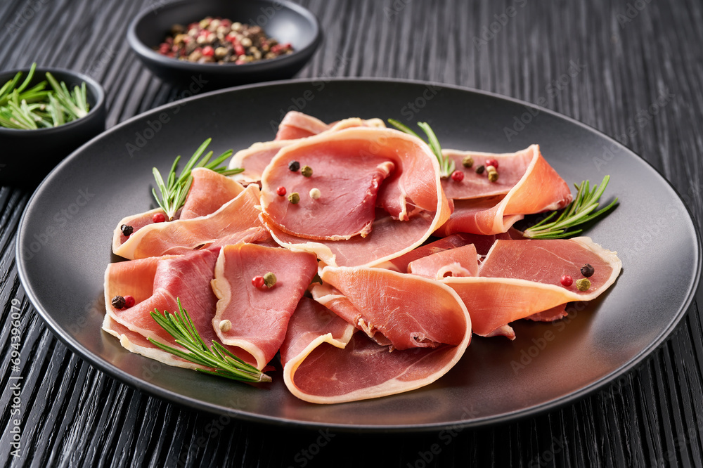 parma ham prosciutto with rosemary and spices in a plate on a black wooden table. parma ham prosciutto with rosemary and spices in a plate on a black wooden table.
