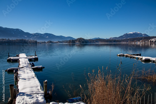 Snow covered wooden pier on lake Faak in Carinthia, Austria, Europe. Surrounded by high snow capped Austrian Alps mountains. Calm water surface with reflections of landscape. Looking at Dobratsch peak photo