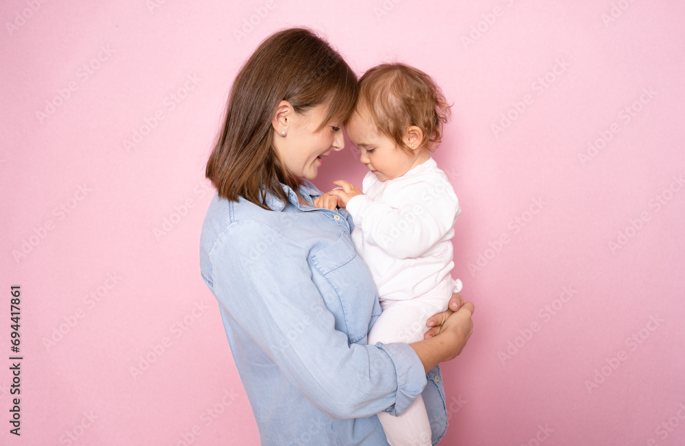 Portrait of satisfied smiling mother holding infant baby girl, happy family having fun together, adorable kid in mom's arms, mom and child isolated over pink background.