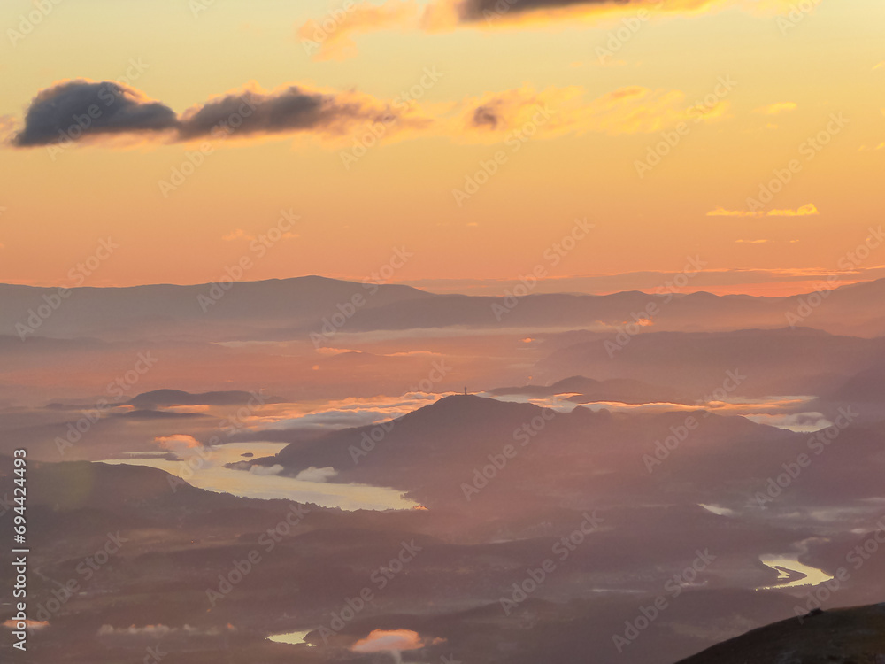 Obraz premium Panoramic sunrise view from Dobratsch on Julian Alps and Karawanks in Austria, Europe. Silhouette of endless mountain ranges. Lake Woerthersee (lake Woerth) surrounded by mysterious haze and fog