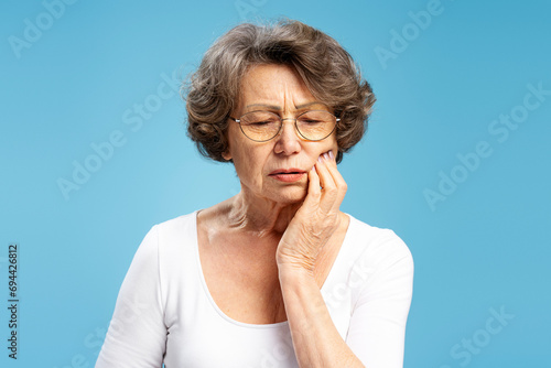 Portrait of upset mature senior  woman wearing eyeglasses touching face having toothache isolated on blue background. Concept of treatment, dental care photo