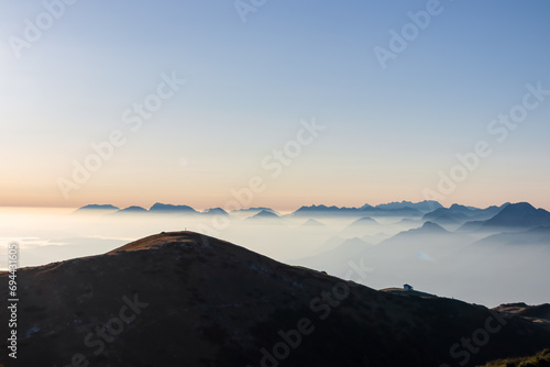 Panoramic sunrise view from Dobratsch on Julian Alps and Karawanks in Austria, Europe. Silhouette of endless mountain ranges with orange and pink sky. Jagged sharp peaks and valleys. Cottage on hill