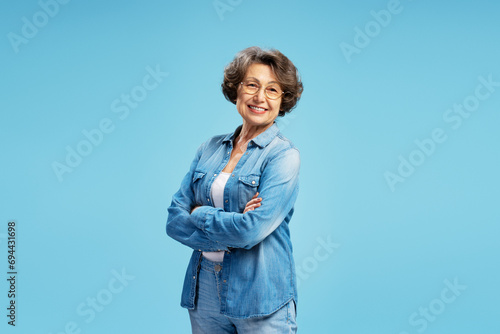 Photography Smiling beautiful senior elderly woman wearing glasses, stylish blue shirt with arms crossed, standing isolated on blue background