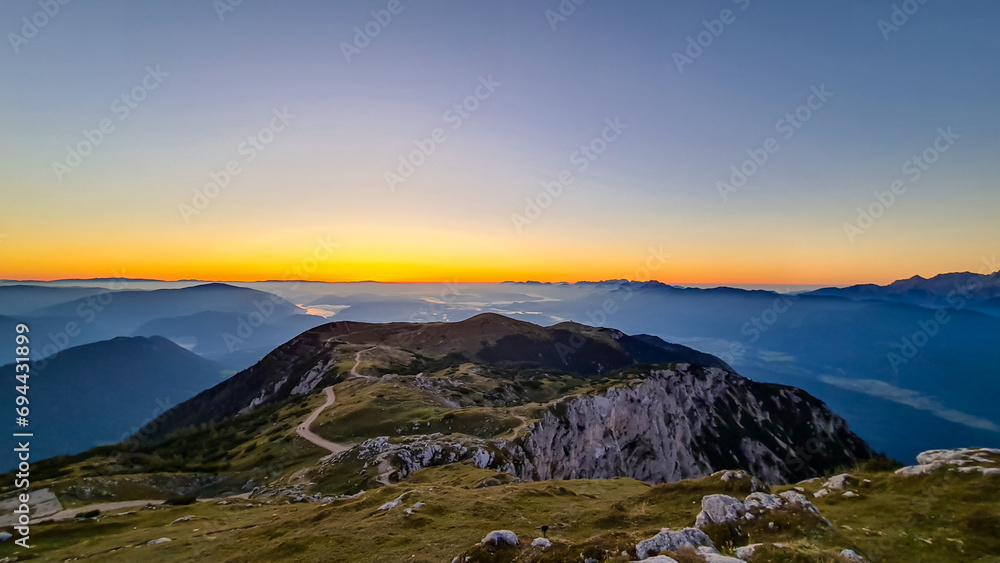 Scenic sunrise view from summit Dobratsch on Julian Alps and Karawanks in Austria, Europe. Silhouette of endless mountain ranges touched by morning sunbeams. Misty fog in valley, tranquil atmosphere