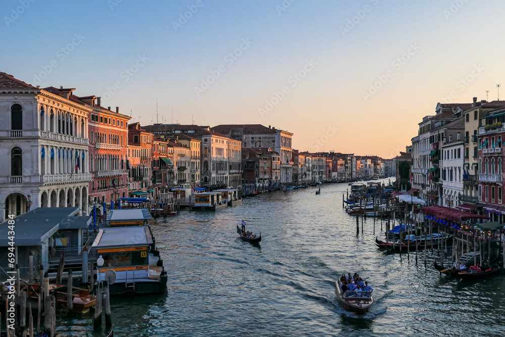 Scenic sunset view of Grand Canal (Grand Canale) with tourist gondolas in Venice, Veneto, Italy, Europe. Famous landmark cathedral San Simeon Piccolo. Gondoliers at golden hour. Urban summer tourism