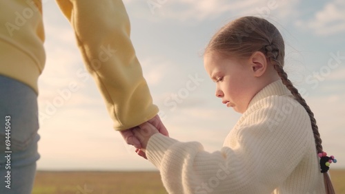 Little daughter, mother, holding hands, are walking. Parent, child, hand in hand. Family walk, daughter, mother. Mother, sad child girl walk together hand in hand in park, sky. Family walk outdoor. photo