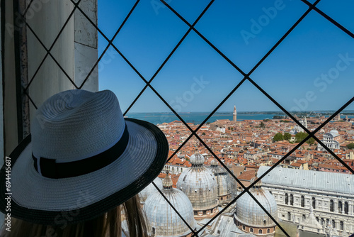 Tourist woman wearing hat with aerial view from St Mark bell tower Campanile of old town Venice, Veneto, Italy, Europe. Looking at Santa Maria Della Salute, Venetian lagoon. UNESCO World Heritage