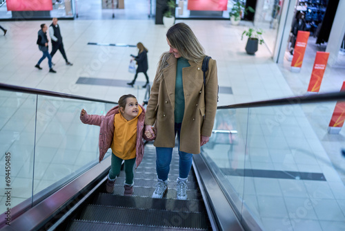 Mother and daughter ascend on the escalator, sharing a moment in the bustling shopping center.