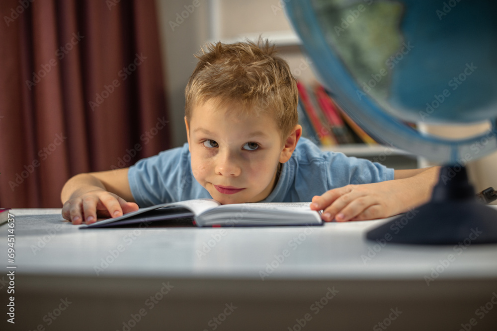 blond boy doing homework at home, tired and looking at the globe on the table blond boy doing homework at home, tired and looking at the globe on the table