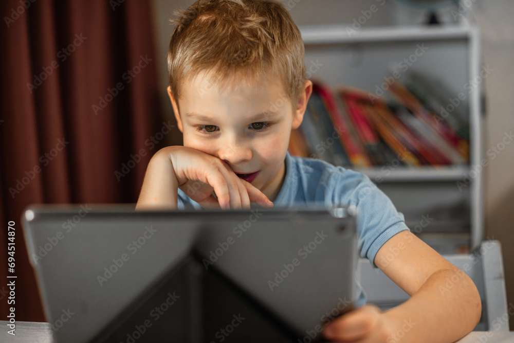A cute blond boy sits in his room at his tablet. smiles and watches the video with pleasure A cute blond boy sits in his room at his tablet. smiles and watches the video with pleasure