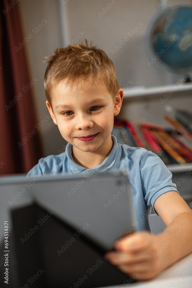 A cute blond boy sits in his room at his tablet. smiles and with interests watches the video with pleasure A cute blond boy sits in his room at his tablet. smiles and with interests watches the video with pleasure