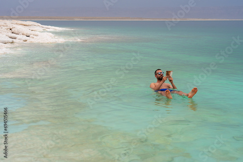 Relaxing tourist man reading a book while taking a bath in floating Death Sea, Jordan