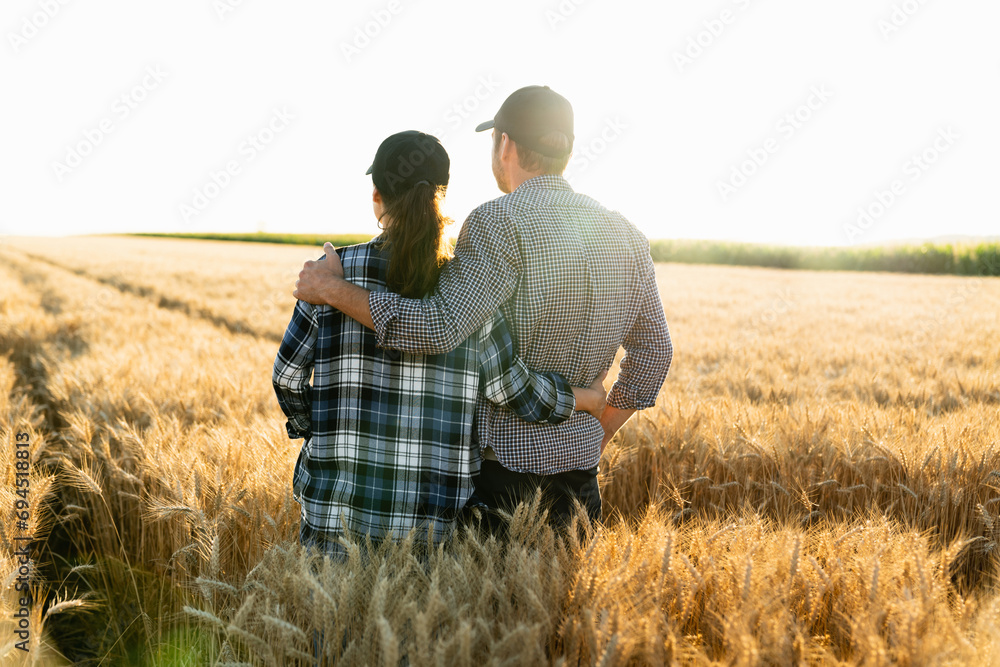 A couple of farmers in plaid shirts and caps stand embracing on agricultural field of wheat at sunset.