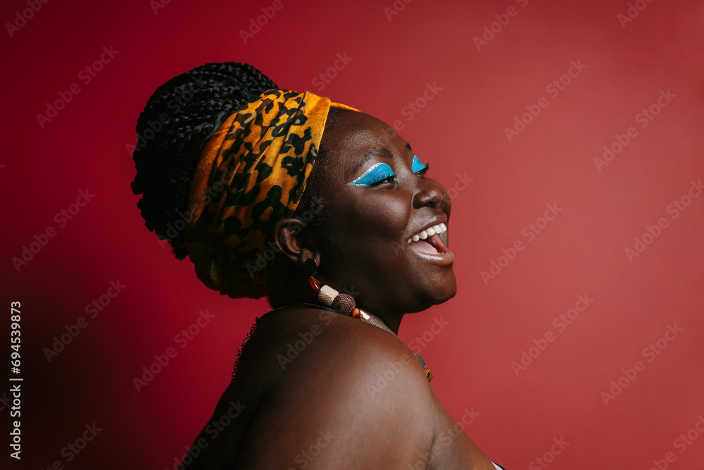Happy plus size African woman with colorful make-up wearing traditional headwear on red background - obrazy, fototapety, plakaty 
