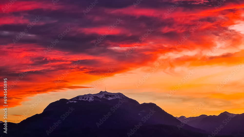 Scenic view of snow capped mountain peak Dobratsch at sunset seen from Taborhoehe in Carinthia, Austria, Europe. Sky has vibrant orange and pink colors with clouds swirling around summit. Serenity