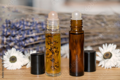 Two liquid cosmetics in roll on transparent and amber glass bottle at wooden background with white flowers and dry lavender bunch.