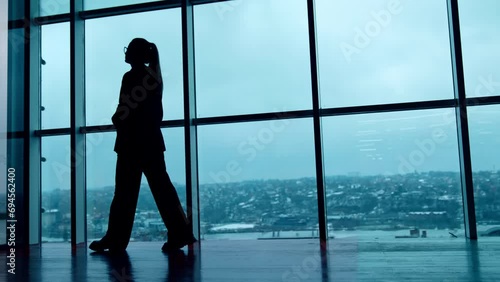 Silhouette of a woman in glasses and suit walks along the panoramic windows. City panorama at backdrop. Low angle view. photo