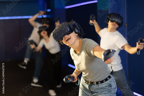 Excited modern young woman with gaming controllers in hands and VR goggles having fun with friends in virtual reality room. Concept of interactive experience..