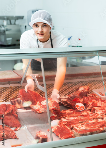 Smiling young butcher working behind counter in butchery, showing fresh raw veal cuts..