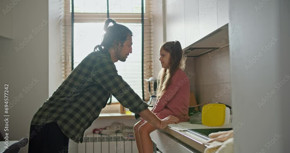 A brunette man in a green checkered shirt leans on the table and communicates with his little brunette daughter in a pink dress in a modern kitchen A brunette man in a green checkered shirt leans on the table and communicates with his little brunette daughter in a pink dress in a modern kitchen