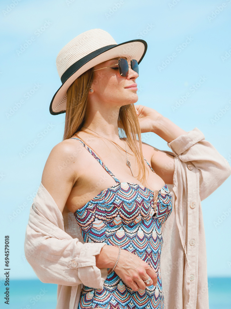 Young woman in a swimsuit, shirt, sunglasses and hat posing gracefully on a sandy beach against a blue sky
