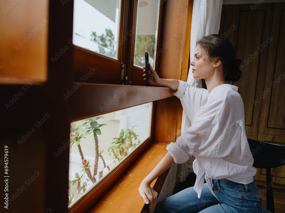 Beautiful woman with phone in hand sitting at the window with wooden frame of the house, home comfortable lifestyle with online work, cozy atmosphere and aesthetics of warmth, spring time Beautiful woman with phone in hand sitting at the window with wooden frame of the house, home comfortable lifestyle with online work, cozy atmosphere and aesthetics of warmth, spring time