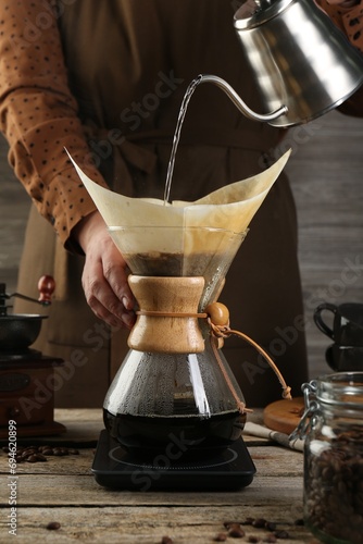 Woman pouring hot water into glass chemex coffeemaker with paper filter at wooden table, closeup
