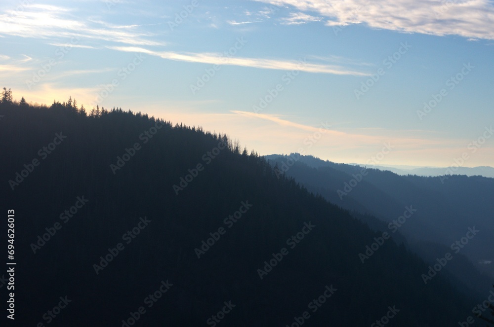 Pine covered mountain range backlit by sunset, with the mountains in shadow on a partially cloudy day near sunset. 