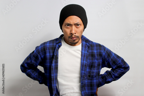 Angry young Asian man, wearing a beanie hat and casual outfit, stands with arms akimbo, looking furious at the camera. Isolated on a white background