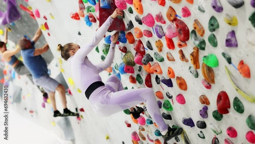 Concentrated young woman in comfortable athletic clothing moving carefully on bouldering wall clinging to colorful hand and foot holds. Popular activity in indoor climbing gym photo