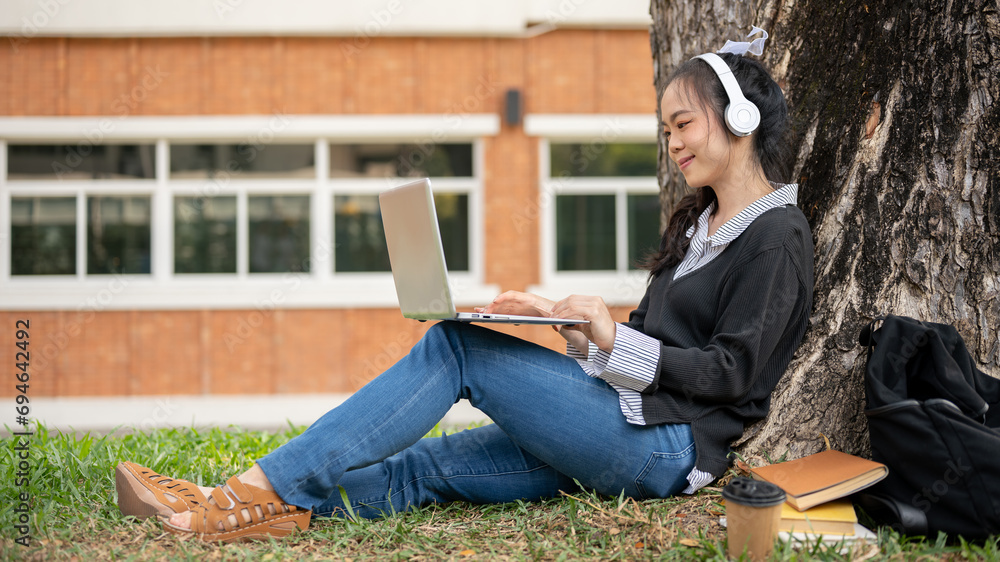 Fototapeta premium A positive young Asian female college student is using her laptop computer under the tree in a park.