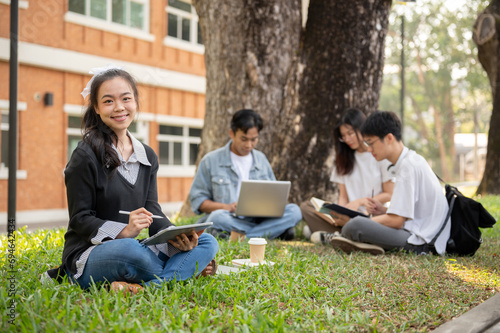 A beautiful Asian female student with her tablet is sitting on grass in a park with her friends.