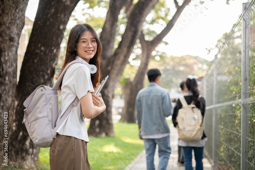 A positive, cute Asian female student with her laptop is standing on a footpath on her campus.