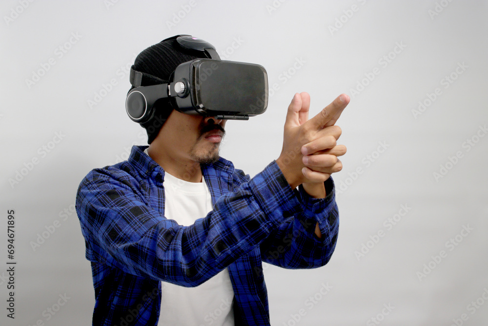Asian man, wearing VR glasses on his head, imitates a finger pistol gesture while engaged in a Virtual Reality shooter game, aiming and shooting at the target while standing against a white background Asian man, wearing VR glasses on his head, imitates a finger pistol gesture while engaged in a Virtual Reality shooter game, aiming and shooting at the target while standing against a white background