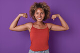 Young strong smiling African American woman teenager looks at screen with smile posing in strongman pose and showing off biceps as sign of self-confidence stands on isolated purple background.