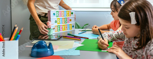 Unrecognizable female teacher holding lightbox with text reduce, reuse, recycle while young students drawing on colorful cardboards in ecology classroom. Sustainable lifestyle education concept. photo