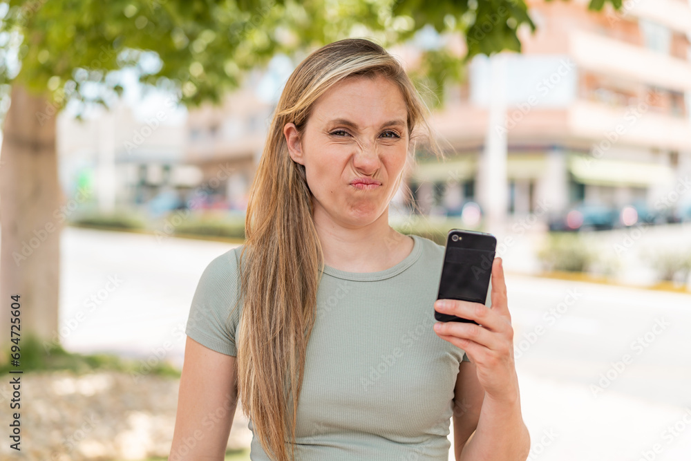 Young blonde woman using mobile phone at outdoors with sad expression Young blonde woman using mobile phone at outdoors with sad expression
