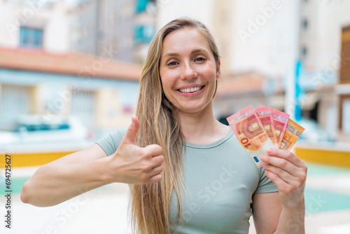 Young blonde woman taking a lot of money at outdoors with thumbs up because something good has happened