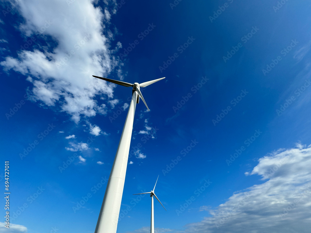 風車と風力発電・大きな海に波が打ち寄せ、青い大空と太陽と夕日が混ざり合い、高台から見下ろす実写写真