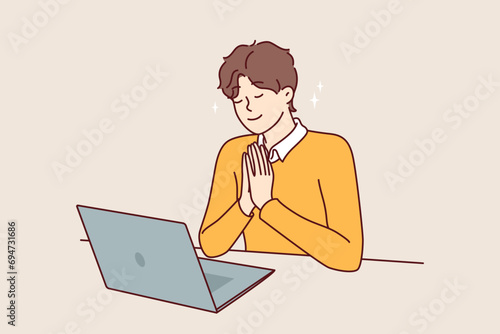Man prays sitting in front of laptop and watching broadcast of religious event from catholic church. Praying young guy believes in god, prays before starting office work with computer. photo