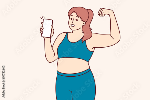 Plump woman in fitness clothes demonstrates biceps and encourages use of mobile phone with sports applications. Girl experiencing problems with excess weight does fitness to become slim and beautiful.