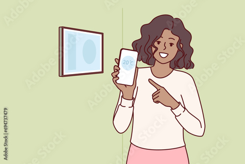 Woman holds phone with thermometer standing near control panel for smart home, and boasts of having iot system. Girl calls to integrate smart home technology for temperature management via smartphone