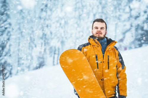 Man At Ski Resort with a snowboard On The snowy Background. Bearded man in winter landscape. Winter holidays in the snowy mountains with active sports.