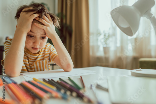 Depressed boy with paper and pencils sitting at desk photo