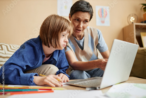 Mother explaining lesson on laptop to daughter with Down syndrome at home photo