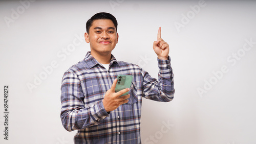 Young Asian man using smartphone over white background with a big smile on face, pointing with hand finger to the side looking at the camera. studio shot photo