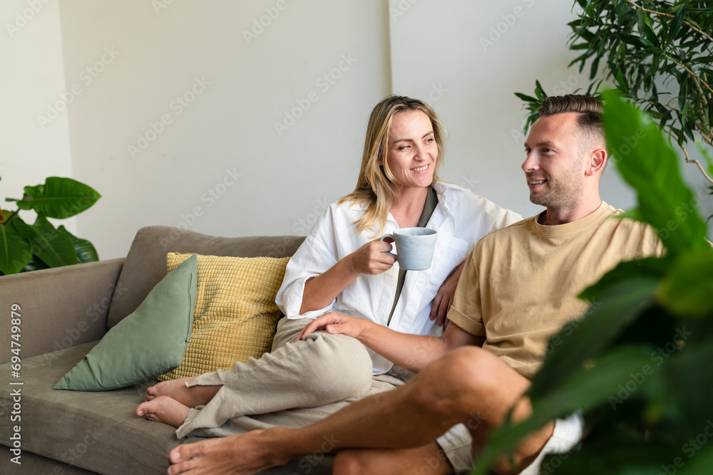 Smiling blond woman with coffee cup talking to man on sofa at home Smiling blond woman with coffee cup talking to man on sofa at home