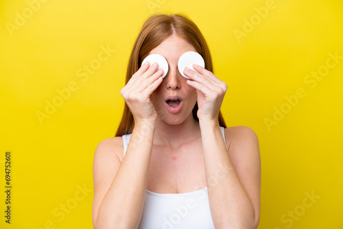 Young redhead woman isolated on yellow background with cotton pad for removing makeup from her face