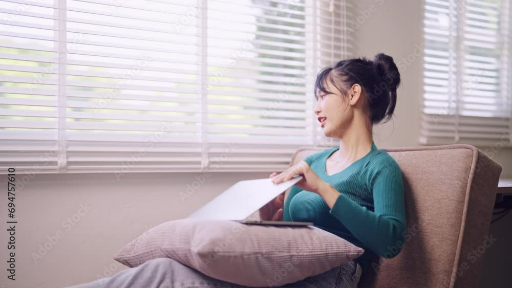 An Asian young woman works on her laptop, taking a comfortable break while seated on her home sofa during the weekend. Capture the serene blend of productivity and relaxation An Asian young woman works on her laptop, taking a comfortable break while seated on her home sofa during the weekend. Capture the serene blend of productivity and relaxation