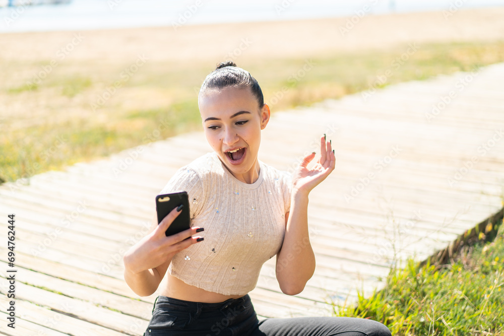 Young moroccan girl  at outdoors looking at the camera while using the mobile with surprised expression